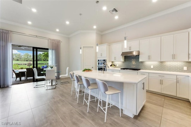 kitchen with under cabinet range hood, visible vents, ornamental molding, light tile patterned floors, and a kitchen island with sink