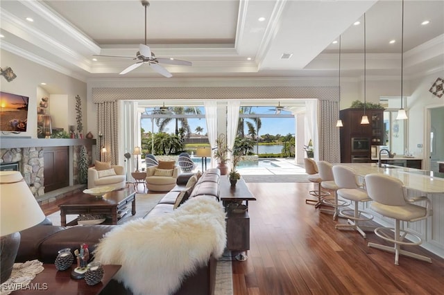 living area with ceiling fan, ornamental molding, a stone fireplace, wood finished floors, and a raised ceiling