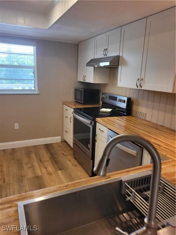 kitchen with under cabinet range hood, baseboards, butcher block counters, white cabinetry, and appliances with stainless steel finishes