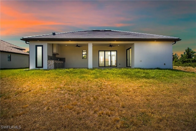 back of house at dusk with a yard, a ceiling fan, and stucco siding