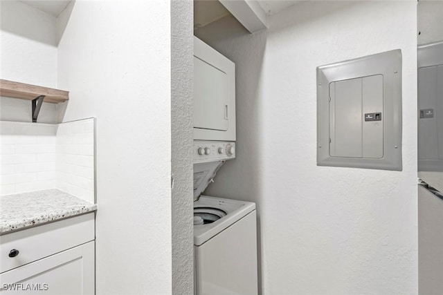 laundry area with electric panel, a textured wall, stacked washer / dryer, and laundry area