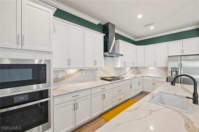 kitchen featuring white cabinets, stainless steel appliances, a sink, wall chimney range hood, and ornamental molding