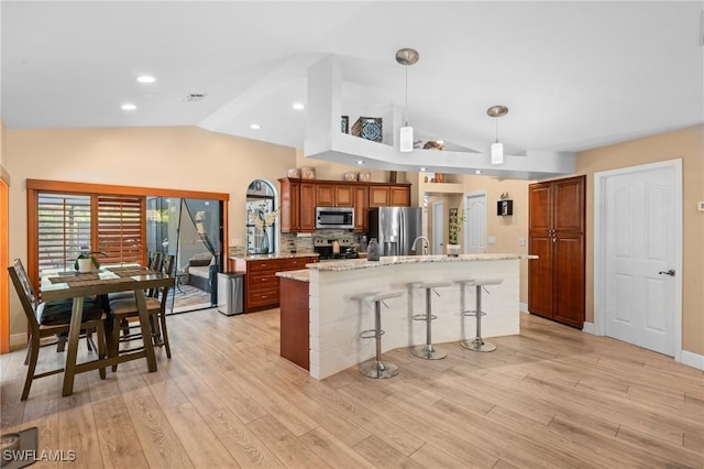 kitchen featuring an island with sink, brown cabinetry, vaulted ceiling, stainless steel appliances, and a breakfast bar area