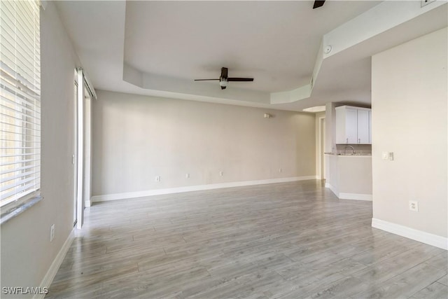 unfurnished living room with a tray ceiling, light wood-style floors, baseboards, and a ceiling fan