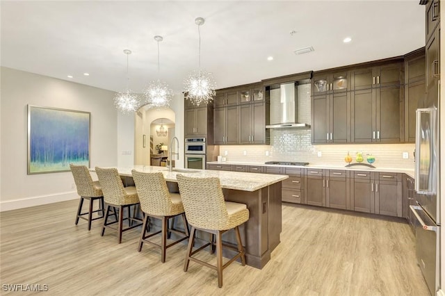 kitchen with wall chimney range hood, tasteful backsplash, light wood-style flooring, stainless steel appliances, and a notable chandelier