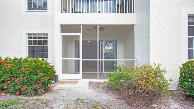 view of exterior entry with stucco siding, ceiling fan, and a balcony