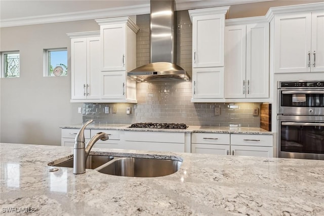 kitchen with white cabinets, a sink, wall chimney range hood, appliances with stainless steel finishes, and crown molding