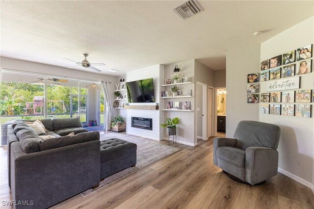 living room featuring ceiling fan, light wood finished floors, a large fireplace, baseboards, and a textured ceiling