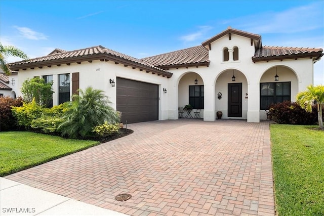 mediterranean / spanish home featuring stucco siding, decorative driveway, a front lawn, a tile roof, and an attached garage