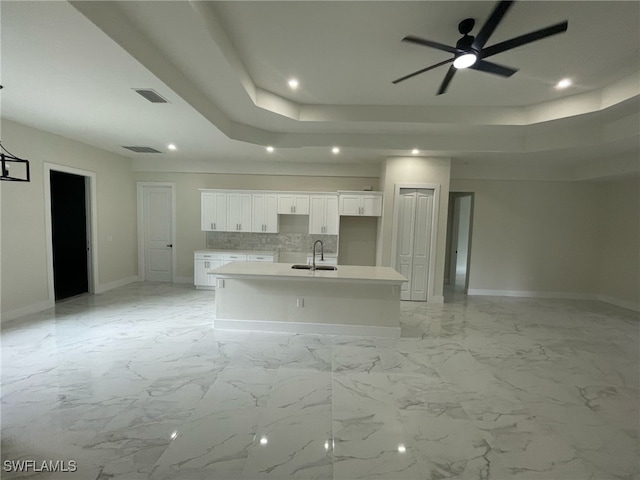 kitchen featuring an island with sink, white cabinetry, a ceiling fan, a tray ceiling, and a sink