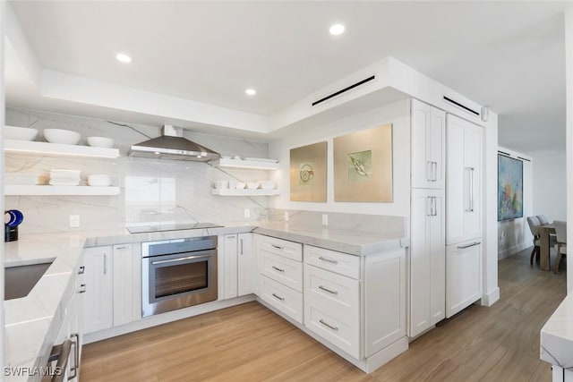 kitchen featuring light wood-style floors, stainless steel oven, wall chimney range hood, open shelves, and black electric stovetop