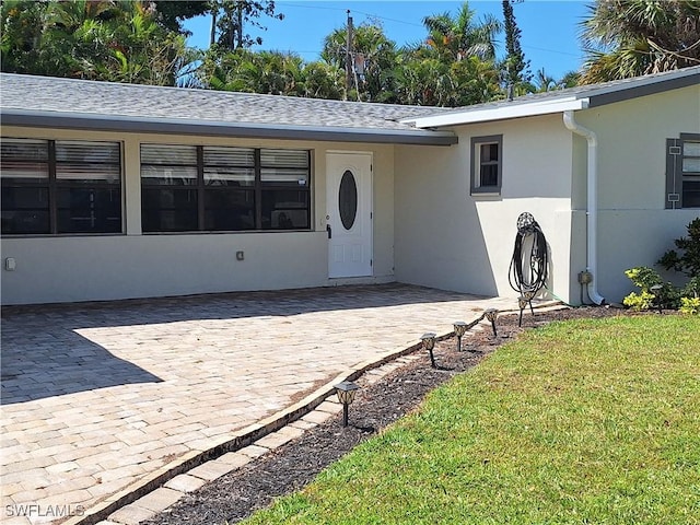 view of exterior entry with roof with shingles, stucco siding, and a lawn