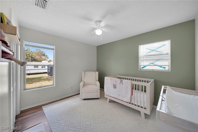 bedroom featuring a nursery area, a ceiling fan, a textured ceiling, wood finished floors, and baseboards