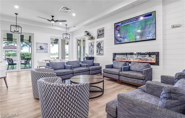 living area with a ceiling fan, a glass covered fireplace, wooden walls, light wood finished floors, and visible vents