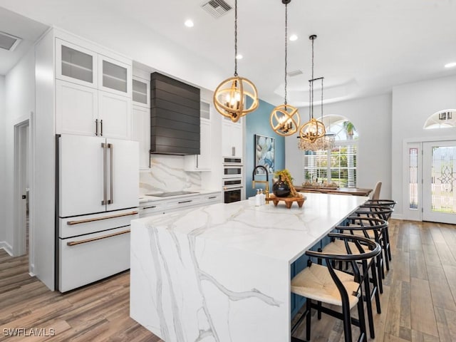 kitchen with white cabinets, a chandelier, wood finished floors, white appliances, and an island with sink