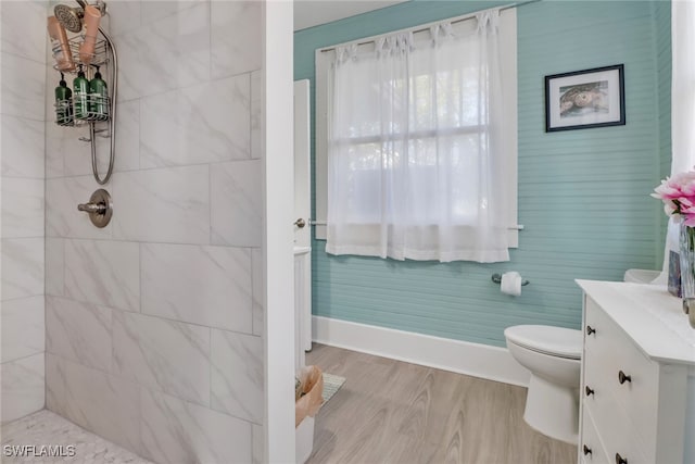 bathroom with toilet, wood finished floors, vanity, a tile shower, and baseboards
