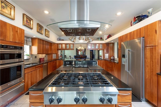 kitchen featuring appliances with stainless steel finishes, brown cabinetry, island range hood, backsplash, and a warming drawer