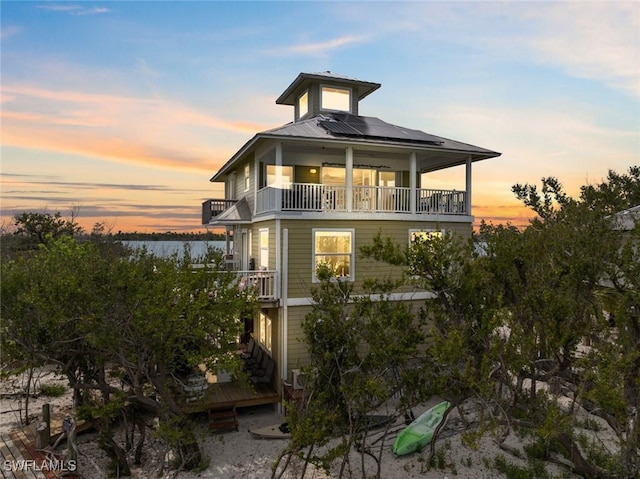 back of house at dusk featuring a water view, roof mounted solar panels, a balcony, and metal roof