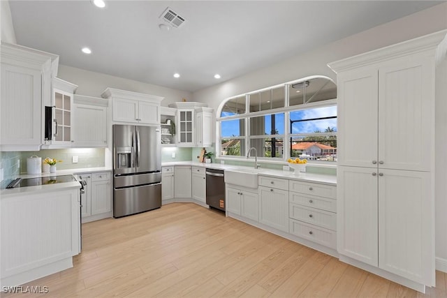 kitchen featuring appliances with stainless steel finishes, a sink, light wood-style flooring, decorative backsplash, and glass insert cabinets