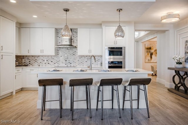 kitchen featuring a sink, a breakfast bar area, white cabinetry, wall chimney range hood, and double oven