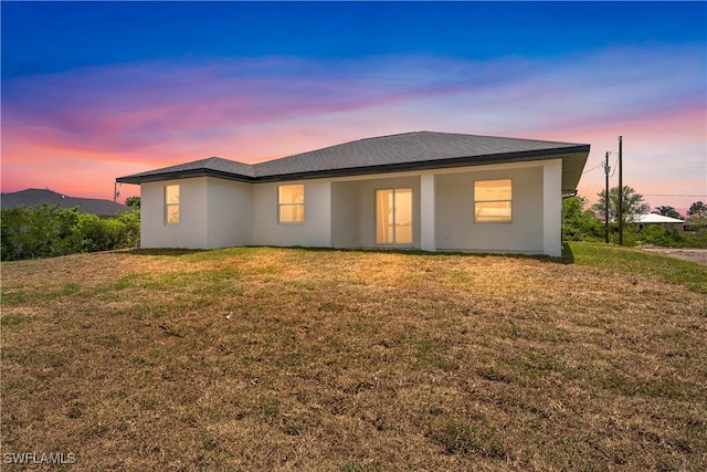 back of house at dusk featuring a lawn and stucco siding