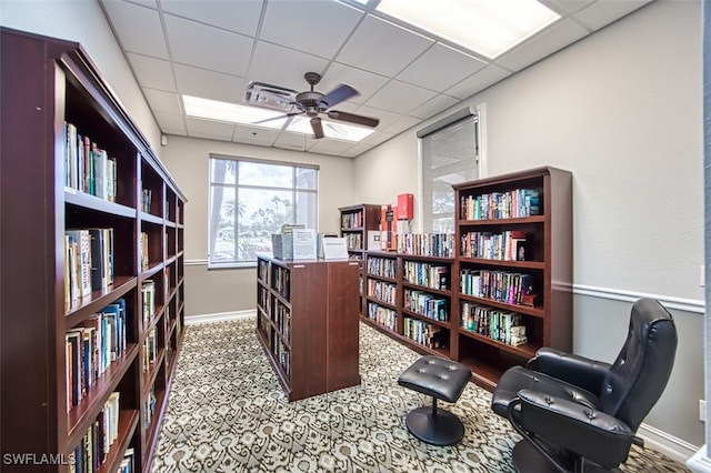 home office featuring a drop ceiling, wall of books, baseboards, carpet flooring, and ceiling fan
