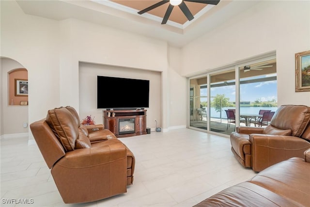 living room with arched walkways, ceiling fan, a tray ceiling, and baseboards