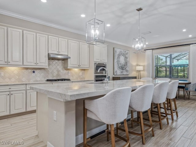 9459 Campanile Cir, Naples, FL 34114 - kitchen featuring under cabinet range hood, gas stovetop, a kitchen island with sink, tasteful backsplash, and ornamental molding