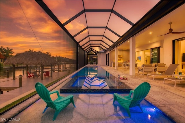 pool at dusk featuring an outdoor pool, a jacuzzi, a patio, ceiling fan, and glass enclosure