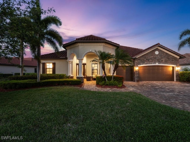 mediterranean / spanish-style home with stucco siding, decorative driveway, stone siding, a front yard, and an attached garage