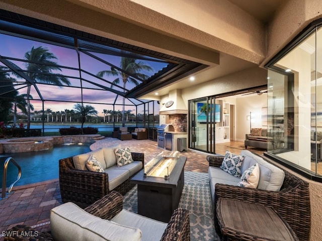 patio terrace at dusk featuring an outdoor living space with a fire pit, exterior kitchen, a water view, glass enclosure, and a pool with connected hot tub