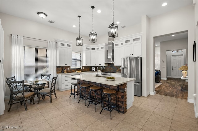 kitchen featuring wall chimney exhaust hood, a center island with sink, backsplash, stainless steel fridge with ice dispenser, and a breakfast bar