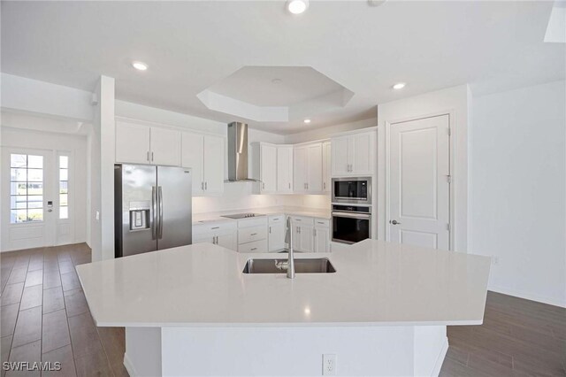 kitchen with wall chimney range hood, dark wood-style flooring, appliances with stainless steel finishes, a sink, and a large island