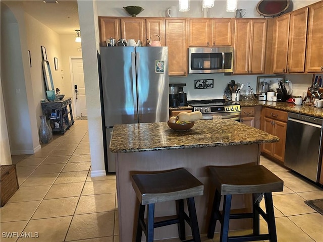 kitchen with stainless steel appliances, light tile patterned floors, a kitchen island, dark stone counters, and a kitchen breakfast bar