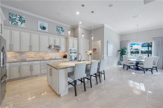 9052 Graphite Cir, Naples, FL 34120 - kitchen featuring under cabinet range hood, backsplash, ornamental molding, stainless steel appliances, and light stone counters