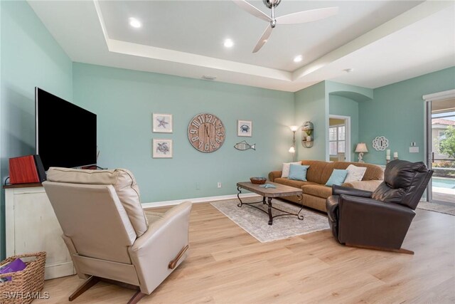 living room featuring light wood-style floors, a ceiling fan, arched walkways, and a tray ceiling