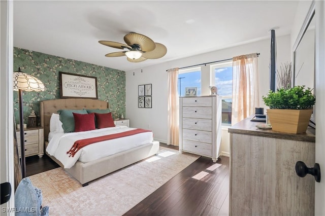 bedroom with an accent wall, ceiling fan, dark wood-style floors, wallpapered walls, and baseboards