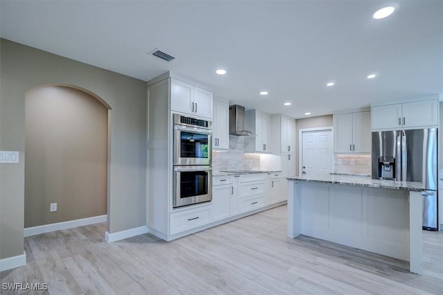 kitchen with wall chimney exhaust hood, light stone counters, arched walkways, backsplash, and stainless steel appliances