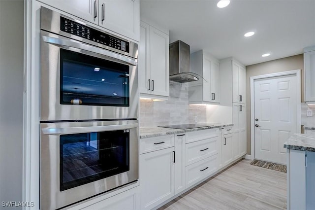 kitchen featuring wall chimney range hood, stainless steel double oven, black electric cooktop, white cabinetry, and light stone counters