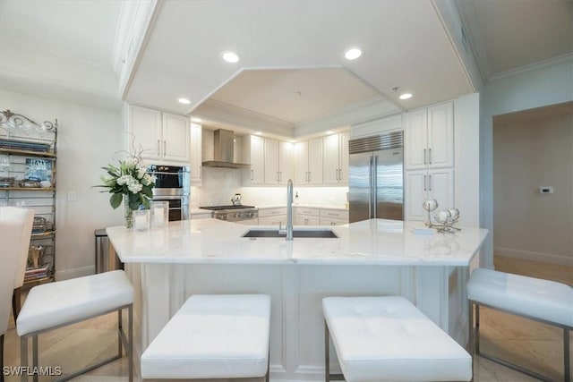 kitchen featuring stainless steel appliances, a sink, wall chimney exhaust hood, ornamental molding, and white cabinetry