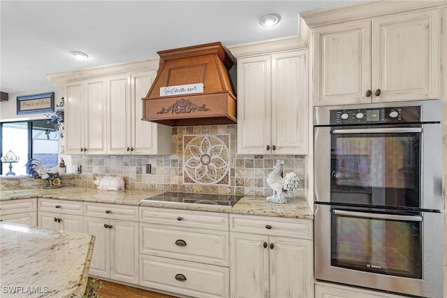 kitchen featuring stainless steel double oven, premium range hood, black electric stovetop, decorative backsplash, and light stone countertops