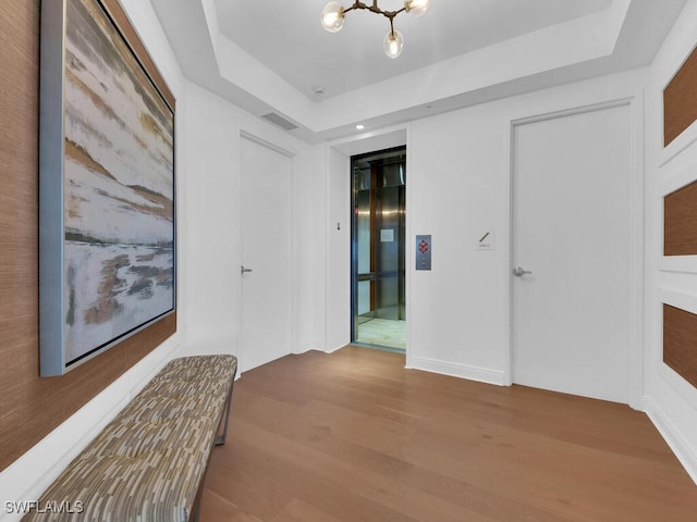 hall with wood finished floors, a chandelier, elevator, and a raised ceiling