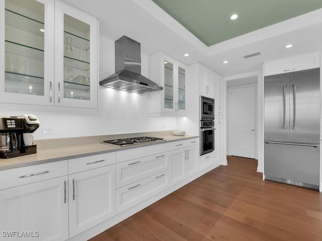 kitchen featuring built in appliances, wall chimney exhaust hood, dark wood-type flooring, white cabinets, and light countertops