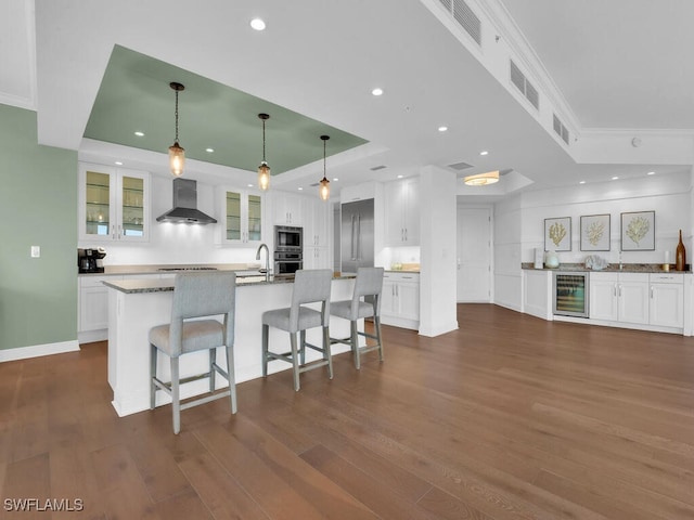 kitchen with a raised ceiling, wall chimney exhaust hood, wine cooler, built in appliances, and white cabinets