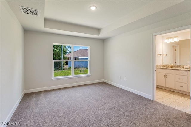 unfurnished bedroom with visible vents, baseboards, light colored carpet, a raised ceiling, and ensuite bath