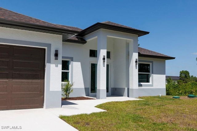 doorway to property featuring stucco siding, an attached garage, a yard, and a shingled roof