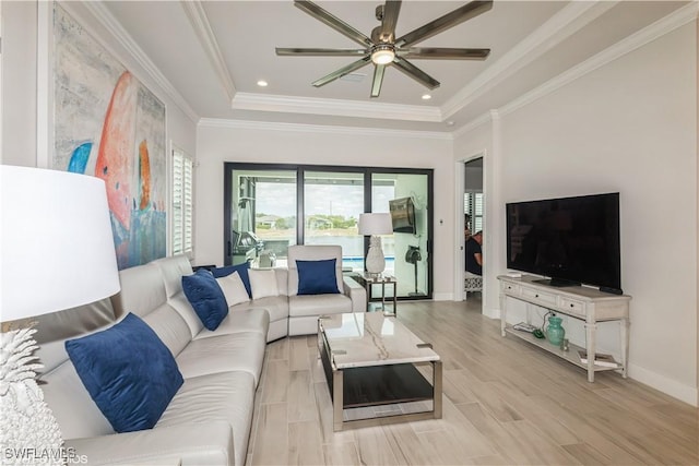 living room featuring a raised ceiling, ceiling fan, baseboards, ornamental molding, and light wood-type flooring