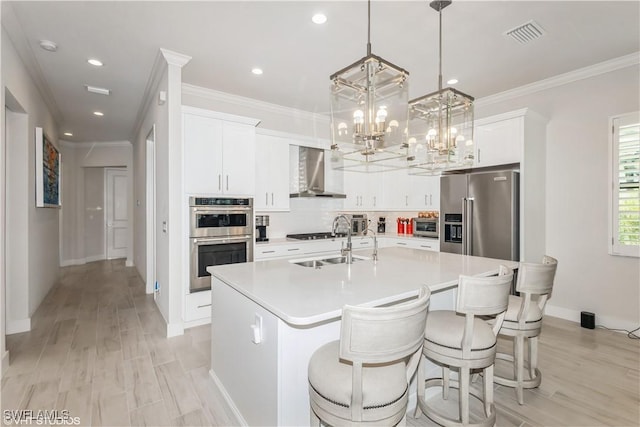 kitchen with wall chimney exhaust hood, stainless steel appliances, a center island with sink, light countertops, and crown molding