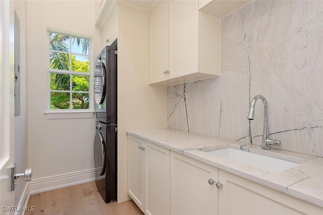 clothes washing area with stacked washer and dryer, cabinet space, a sink, baseboards, and light wood finished floors