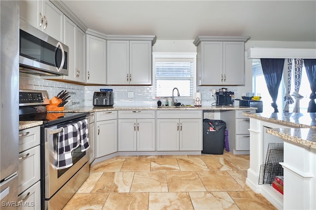 kitchen with stainless steel appliances, a sink, decorative backsplash, gray cabinetry, and light stone counters